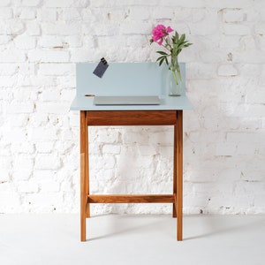 Może przedstawiać: Jasnoniebieskie biurko z drewnianą ramą i jedną szufladą. Biurko ma małą półkę nad szufladą. Laptop znajduje się na biurku. Wazon z różowymi kwiatami stoi na biurku.