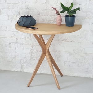 Può includere: Un tavolo console semicircolare in legno con finitura naturale chiara e quattro gambe incrociate. Sul piano del tavolo si trovano una ciotola in feltro grigio scuro piegato, un piccolo smartphone, un vaso rosa testurizzato e una pianta in un vaso blu scuro.
