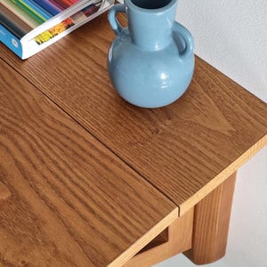 May include: A light blue ceramic vase with two side handles sits on a wooden table with a visible grain pattern. Next to the vase, a book with a colorful striped cover is partially visible, showing the text Kassia St. Clair on the spine.
