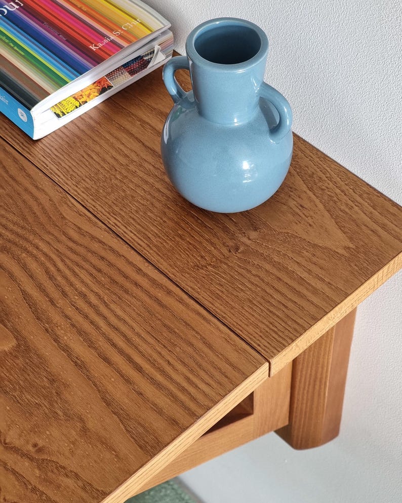 May include: A light blue ceramic vase with two side handles sits on a wooden table with a visible grain pattern. Next to the vase, a book with a colorful striped cover is partially visible, showing the text Kassia St. Clair on the spine.
