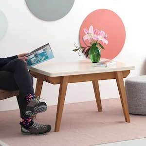 Könnte beinhalten: Ein quadratischer Couchtisch mit weißer Platte und hellbraunen Holzbeinen. Eine grüne Glasvase mit rosa Lilien steht auf dem Tisch. Der Tisch ist etwa 60 cm lang und breit und 45 cm hoch.