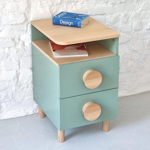 Può includere: Un comodino verde chiaro con due cassetti e un ripiano superiore. I cassetti hanno pomelli rotondi in legno. Il comodino ha un piano in legno chiaro e poggia su quattro gambe in legno.