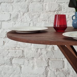 May include: A wooden table with a round top and a white plate and a red glass on the surface. The table is in front of a white brick wall.
