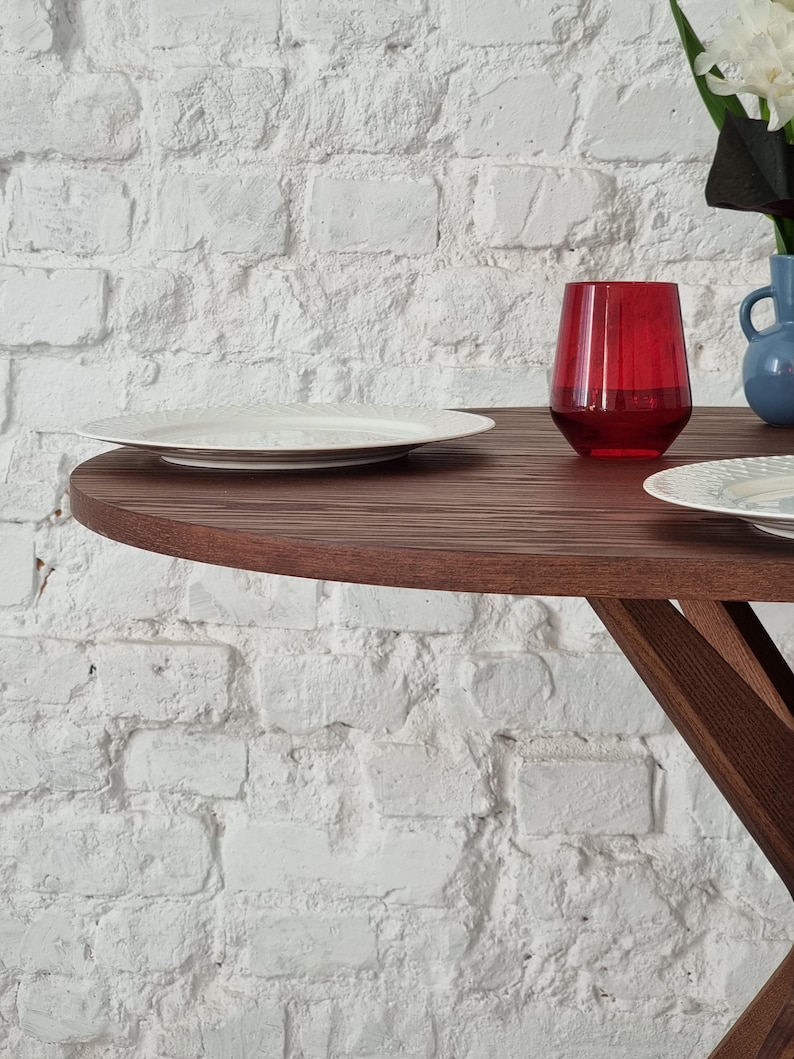 May include: A wooden table with a round top and a white plate and a red glass on the surface. The table is in front of a white brick wall.