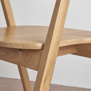 May include: A close-up of a wooden chair with a curved seat and angled legs. The chair is made of light-colored wood and has a natural finish.