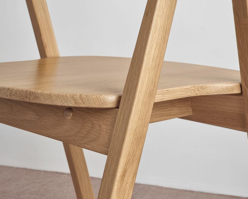 May include: A close-up of a wooden chair with a curved seat and angled legs. The chair is made of light-colored wood and has a natural finish.