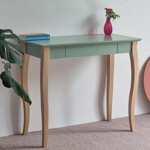 Op de afbeelding: Een saliegroene consoletafel met een glad rechthoekig blad en een enkele lade. De tafel heeft vier lichtgekleurde houten poten met een gebogen ontwerp. Een blauw boek ligt aan de linkerkant en een magenta glazen vaas met een bloem staat rechts.