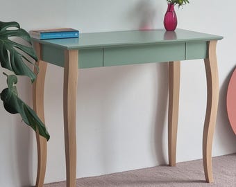 Puede incluir: Una mesa consola de color verde salvia con una superficie rectangular lisa y un cajón. La mesa cuenta con cuatro patas de madera clara con un diseño curvado. Un libro azul descansa a la izquierda y un jarrón de cristal magenta con una flor a la derecha.