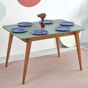 Może przedstawiać: Prostokątny stół jadalny z jasnozielonym blatem i drewnianymi nogami. Na stole ustawiono sześć niebieskich talerzy oraz mały wazon z różowymi kwiatami. Stół stoi na różowym dywanie.