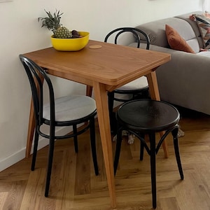 May include: A wooden table with a light brown finish and a black metal frame. The table is set with two black chairs and a black stool. The chairs have white cushions. A yellow bowl with fruit sits on the table.