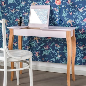 May include: A light pink vanity table with a flip-top mirror, light wood curved legs, and a white wooden chair. On the table surface are two small dark bottles and a blue glass vase containing a single pink flower.