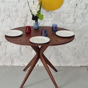 Może przedstawiać: Okrągły drewniany stół z ciemnobrązowym wykończeniem i czterema nogami. Stół jest nakryty trzema białymi talerzami, dwoma niebieskimi szklankami i jedną czerwoną szklanką. Wazon z białymi kwiatami znajduje się w środku stołu.