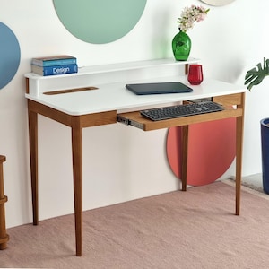 Może przedstawiać: Nowoczesne białe biurko z wysuwaną szufladą na klawiaturę i półką. Biurko ma drewniane nogi i biały blat. Na biurku znajduje się laptop i klawiatura. Zielony wazon i czerwone szkło stoją na półce. Biurko znajduje się w pokoju z różowym dywanem.