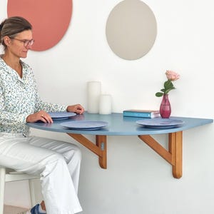 May include: A light blue, half-circle wall-mounted table with wooden supports. Two blue plates sit on the table. A pink rose in a small vase and two white candles are also on the table. The table is approximately 36 inches wide.