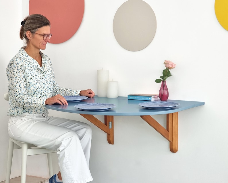 May include: A light blue, half-circle wall-mounted table with wooden supports. Two blue plates sit on the table. A pink rose in a small vase and two white candles are also on the table. The table is approximately 36 inches wide.