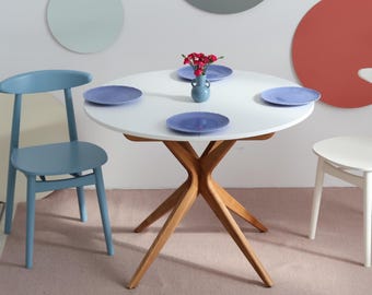 May include: A white circular dining table with a wooden base is set with four blue plates and a small vase of red flowers. A blue chair and a white chair are positioned nearby. Decorative circles in various colours adorn the wall.