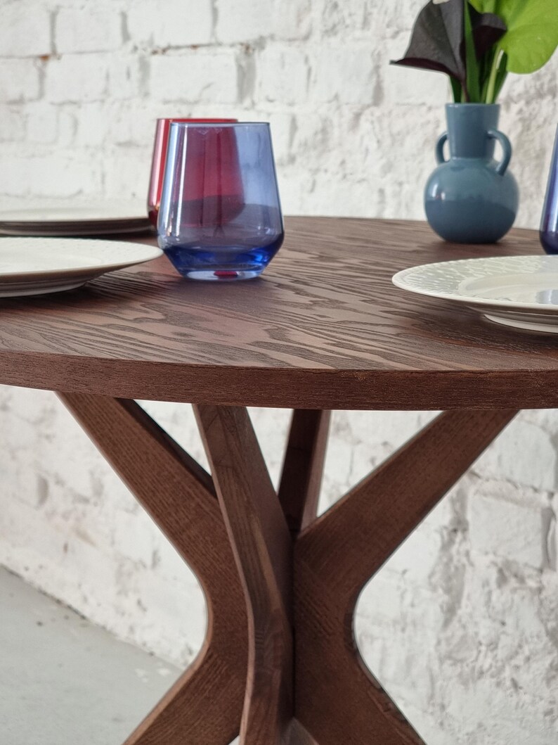 May include: A round wooden table with a dark brown finish and a unique X-shaped base. Two clear glasses with blue and red hues are on the table, along with two white plates.