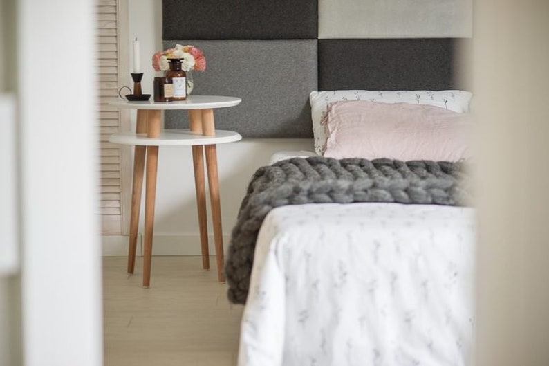 Puede incluir: Una mesita de noche blanca de dos niveles con patas de madera. La mesa est&aacute; junto a una cama con una manta de punto gris y una almohada rosa.