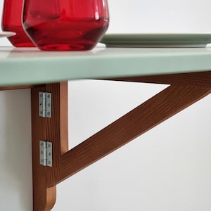 May include: A wall-mounted folding table with a light green surface and a brown wooden support structure. Two red glasses and a green plate are on the table. The table is attached to the wall with metal hinges.