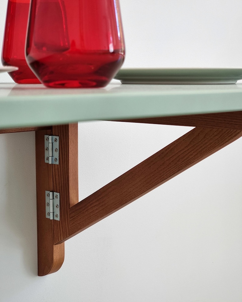 May include: A wall-mounted folding table with a light green surface and a brown wooden support structure. Two red glasses and a green plate are on the table. The table is attached to the wall with metal hinges.