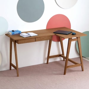 Op de afbeelding: Een L-vormig houten bureau met een natuurlijke bruine afwerking, voorzien van twee geïntegreerde lades aan de linkerkant en een onderstel in schraagstijl. Een blauw boek met de titel Design, een open notitieboek en een laptop liggen op het blad.