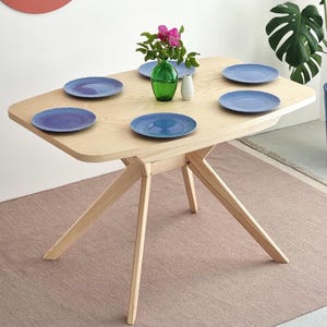 Może przedstawiać: Jasny drewniany stół jadalny z sześcioma niebieskimi talerzami, zielonym wazonem z różowymi kwiatami i białym wazonem. Stół ma unikalny kształt nóg i stoi na różowym dywanie. Stół ma około 122 cm długości.