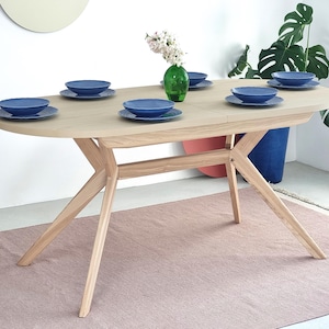 Op de afbeelding: Een ovale houten eettafel met zes blauwe kommen en borden. De tafel heeft een lichte houtafwerking en een uniek pootontwerp. Een groene vaas met bloemen staat in het midden. De tafel staat op een roze tapijt. De tafel is ongeveer 180 cm lang.