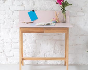 May include: A light pink desk with two natural wood drawers and four slender wooden legs. On the desk surface sits an open book, a small colorful owl figurine, a wooden rabbit cutout, and a glass vase containing a single pink peony flower.