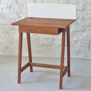 Op de afbeelding: Een klein houten bureau met een enkele lade en een witte achterwand. Het bureau heeft een donkerbruine houten afwerking en een eenvoudig ontwerp.