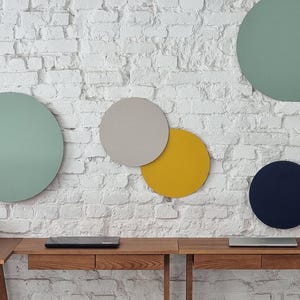 May include: A white brick wall with five round acoustic panels in different colours: green, grey, yellow, and blue. The panels are mounted on the wall above a wooden desk with two drawers.