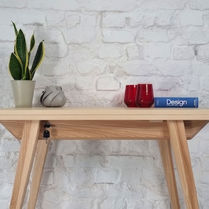May include: A light wood console table with a white brick wall background. The table has a keyhole and a key in the lock. There are two red glasses and a blue book with the word "Design" on the cover on the table.
