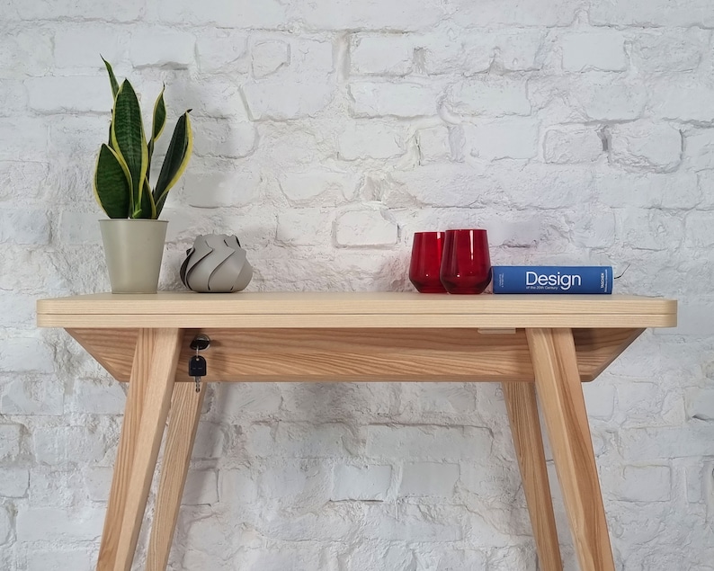 May include: A light wood console table with a white brick wall background. The table has a keyhole and a key in the lock. There are two red glasses and a blue book with the word "Design" on the cover on the table.