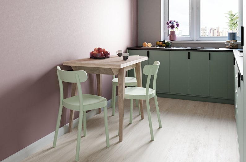 May include: A light green wooden dining table with four light green chairs. The table is set with a bowl of fruit and two mugs. The table is in a kitchen with light green cabinets and a pink wall.