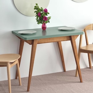 Op de afbeelding: Een retro eettafel met een teal blad en houten poten. Twee witte borden staan op de tafel, met een vaas met roze bloemen. Een houten stoel en kruk zijn ook zichtbaar. De tafel is ongeveer 120 cm lang.