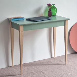Op de afbeelding: Een saliegroen bureau met vier lichtgekleurde houten poten en een lade. Op het bureaublad staan een gesloten zwarte laptop, een boek met een blauwe kaft, een kleine paarse glazen vaas met bloemen en een grote groene glazen fles.