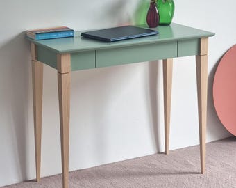 May include: A sage green desk with four light-colored wooden legs and a single drawer. On the desk surface sits a closed black laptop, a book with a blue cover, a small purple glass vase with flowers, and a large green glass bottle.