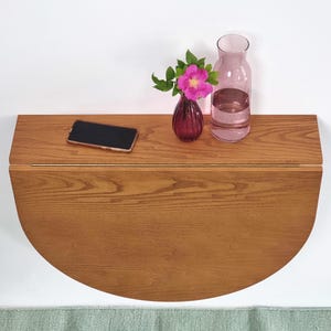 May include: A wall-mounted, semi-circular wooden drop-leaf table with a natural grain finish. On the surface sits a black smartphone, a small dark purple glass vase containing a single pink flower, and a clear glass carafe filled with water.