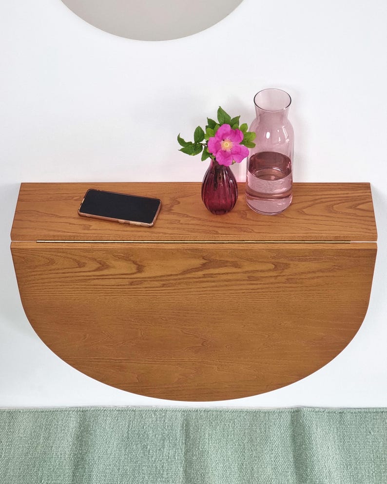 May include: A wall-mounted, semi-circular wooden drop-leaf table with a natural grain finish. On the surface sits a black smartphone, a small dark purple glass vase containing a single pink flower, and a clear glass carafe filled with water.