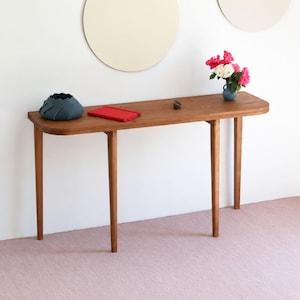 Peut inclure: Une console en bois longue et étroite avec des bords arrondis et quatre pieds fuselés. Sur le dessus se trouvent un bol texturé bleu foncé, un carnet rectangulaire rouge et un petit vase bleu contenant des fleurs roses et blanches.