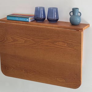 May include: A wooden wall-mounted fold-down table with a light brown finish. The table has a shelf with a book titled "The World Atlas of Coffee" and two blue glass cups. A blue ceramic vase is on the shelf.