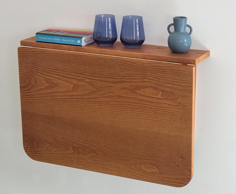 May include: A wooden wall-mounted fold-down table with a light brown finish. The table has a shelf with a book titled "The World Atlas of Coffee" and two blue glass cups. A blue ceramic vase is on the shelf.