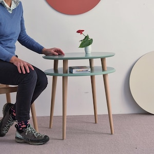 Peut inclure: Une table d'appoint à deux niveaux avec des surfaces ovales vert clair et quatre pieds fuselés en bois clair. La table présente un petit vase blanc avec une fleur rouge et un livre posé sur l'étagère inférieure, sur un sol clair.