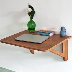 Może przedstawiać: Drewniane biurko montowane na ścianie z laptopem, książką i dwoma wazonami z kwiatami. Biurko wykonane jest z jasnobrązowego drewna i ma zakrzywiony brzeg. Laptop jest srebrny i ma czarną klawiaturę. Książka jest niebieska i ma tęczową okładkę. Wazy są zielone i niebieskie i trzymają białe kwiaty.