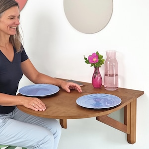 Może przedstawiać: Półokrągły, montowany na ścianie stół z drewna o naturalnym wykończeniu. Na stole znajdują się dwa niebieskie talerze. Na stole stoi różowy wazon z kwiatem i różowa karafka na wodę. Stół jest zamontowany na białej ścianie.