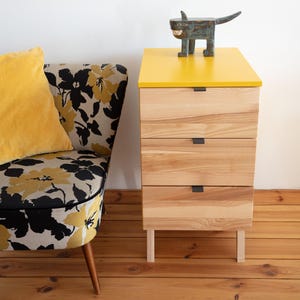 Peut inclure: Une commode en bois à trois tiroirs avec un plateau supérieur jaune vif, dotée de façades en bois clair et de petites poignées noires. Une figurine d'animal bleu foncé aux dents pointues est posée sur le dessus.