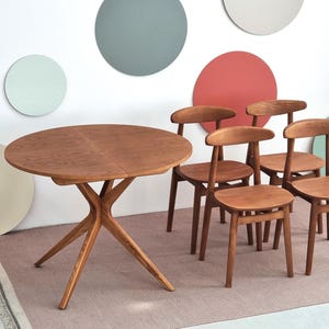 Può includere: Un set da pranzo in legno con un tavolo rotondo e diverse sedie. Il tavolo ha una base scultorea unica e una finitura marrone chiaro. Le sedie hanno schienali curvi e una tonalità di legno simile. Il tavolo ha un diametro di circa 122 cm.