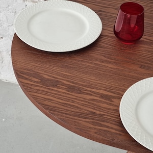 May include: A round wooden table with a dark brown wood grain finish. Two white plates with a decorative pattern are on the table, along with a red glass.