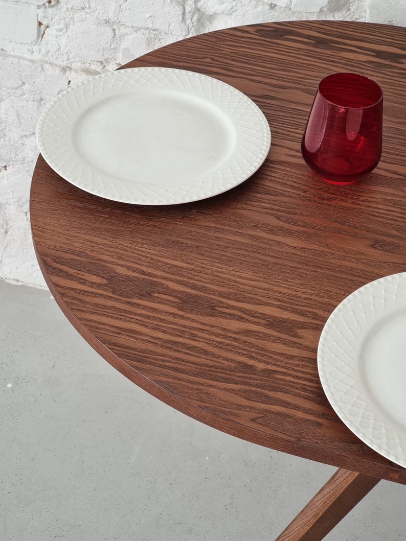 May include: A round wooden table with a dark brown wood grain finish. Two white plates with a decorative pattern are on the table, along with a red glass.