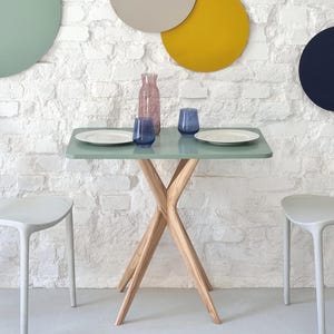 Może przedstawiać: Kwadratowy stół w jasnozielonym kolorze z drewnianym, krzyżowym stelażem. Na stole znajdują się dwa białe talerze i dwa niebieskie szklanki. Stół stoi przed białą ceglaną ścianą z kolorowymi kołami.