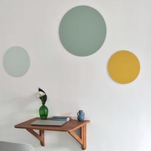 May include: Three round wall decorations in a minimalist home office setting. The largest circle is a light green colour, the medium circle is a light blue colour, and the smallest circle is a yellow colour. A small wooden folding desk with a laptop, a vase with a white flower, and a blue ceramic vase is in the foreground.